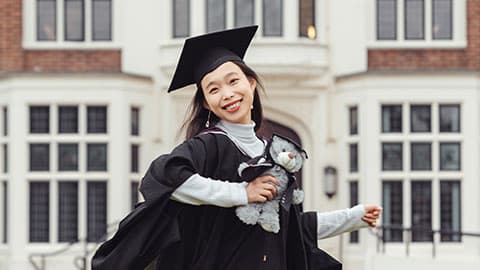 Xu wearing a graduation gown and cap holding her degree scroll outside Hazlerigg Building on Loughborough University campus. She is smiling and she is also holding a grey stuffed teddy bear which is also wearing a black cap and gown.